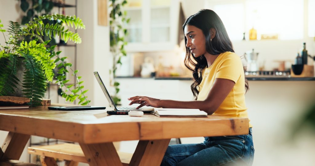 Female student typing on a laptop at a wooden kitchen table for an online course